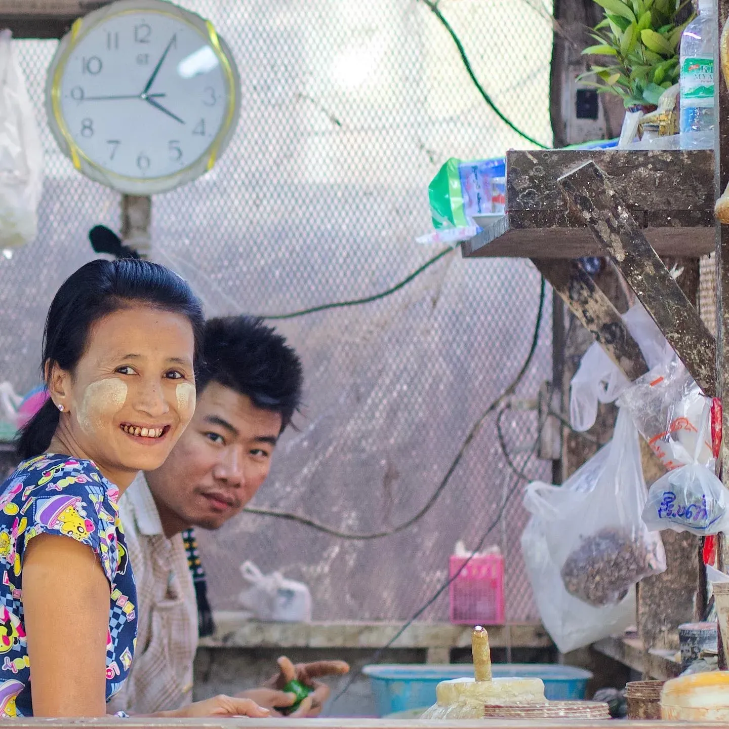 Junge Frau mit Thanaka im Gesicht lacht an einem Marktstand, ein junger Mann schaut hinter ihr hervor, darüber eine Wanduhr